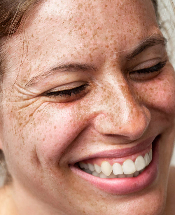 Close-up of a smiling woman with freckles and her eyes closed, highlighting her natural complexion.